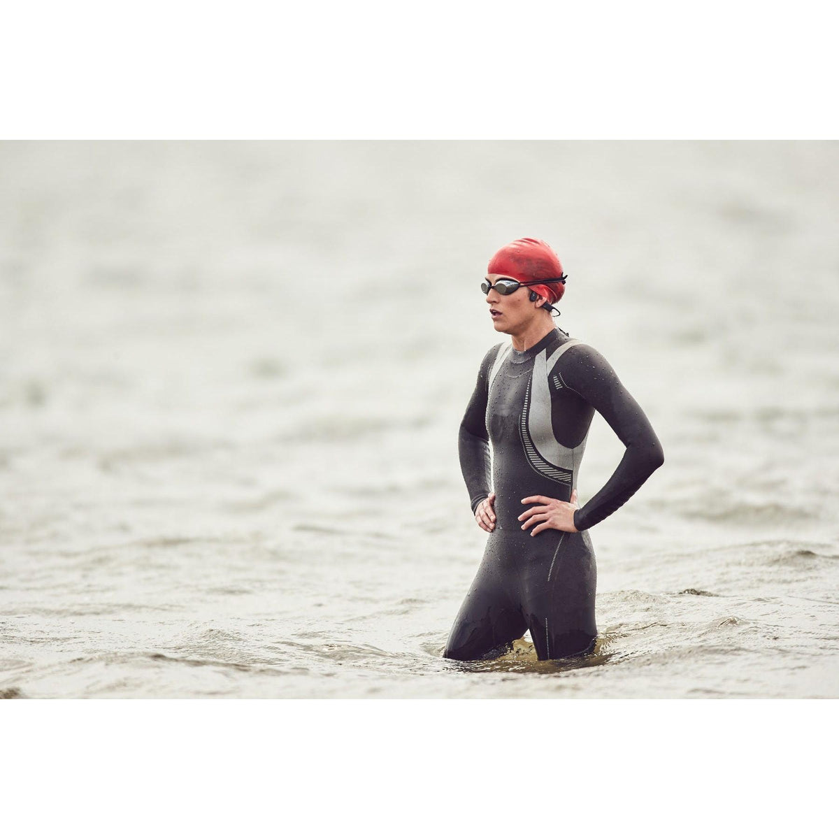 Swimmer wearing black Aftershokz Xtrainerz bone conduction headphones, a wetsuit, and a red swim cap in open water.