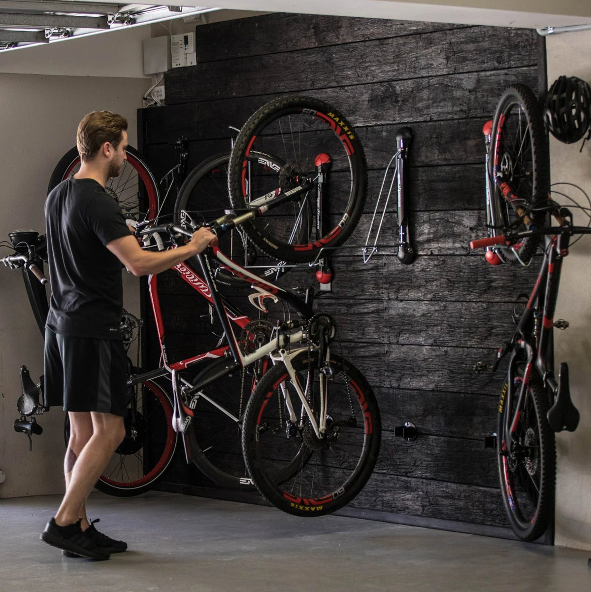 A man in a black t-shirt and shorts is holding a red and black mountain bike in a garage setting with a dark wood-paneled wall holding several other bikes.